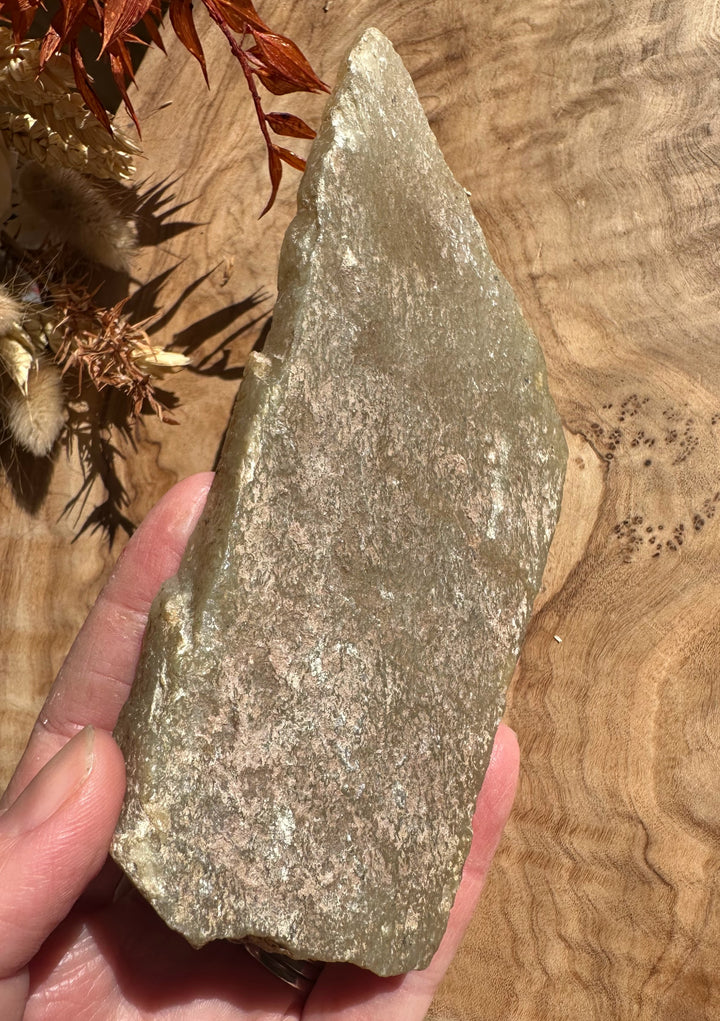 Greenish gray rough jade shard with a jagged surface held above a wooden background