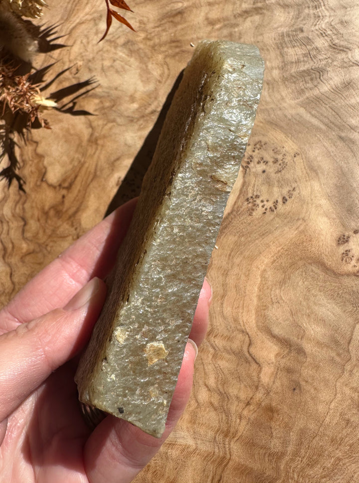 Long narrow rough jade piece with gray green color held in hand against a wood background