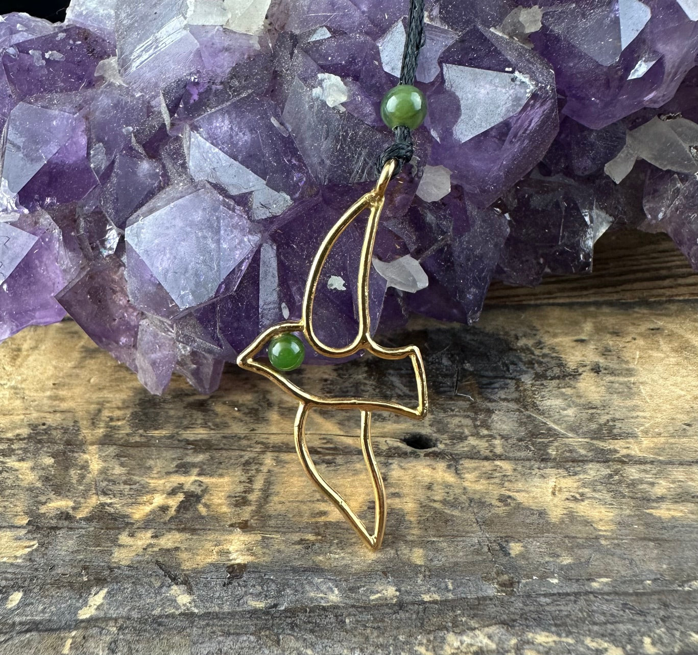 Gold brass bird pendant with two green jade beads against a purple amethyst backdrop.