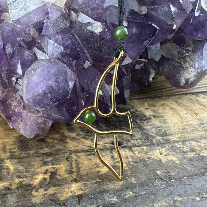 Gold brass bird pendant with two green jade beads displayed on a rustic wood surface with amethyst cluster.
