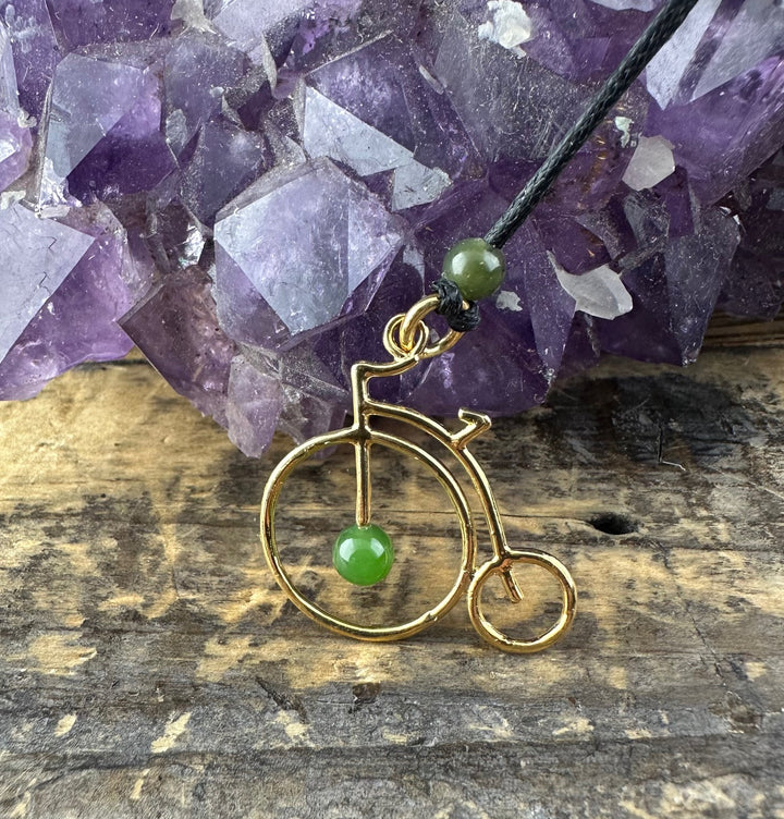 Gold brass penny farthing pendant with a green jade bead, attached to a black cord beside a purple amethyst cluster.