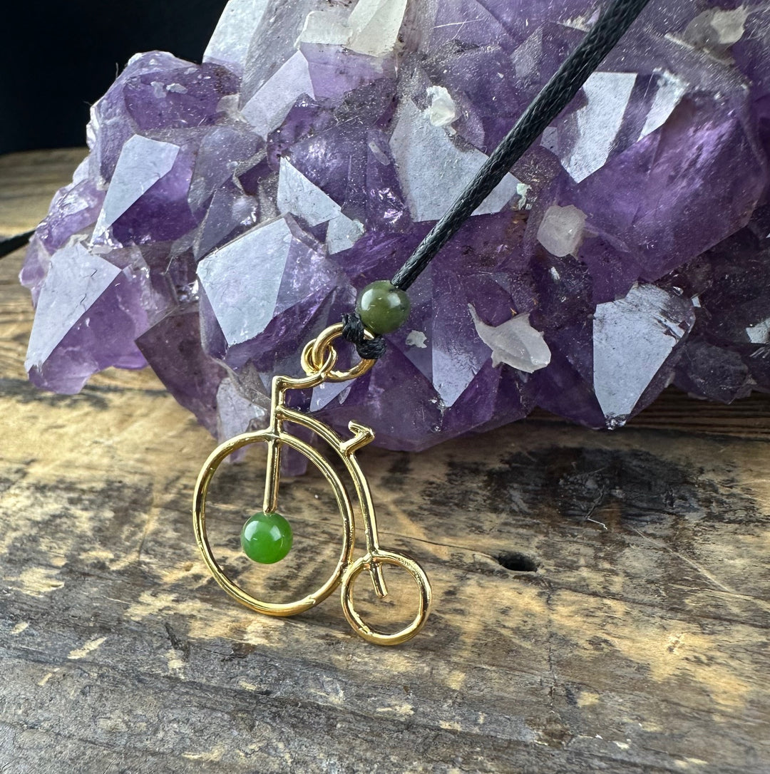 Close up of the brass penny farthing pendant with a green jade bead on a black cord against a purple amethyst backdrop.