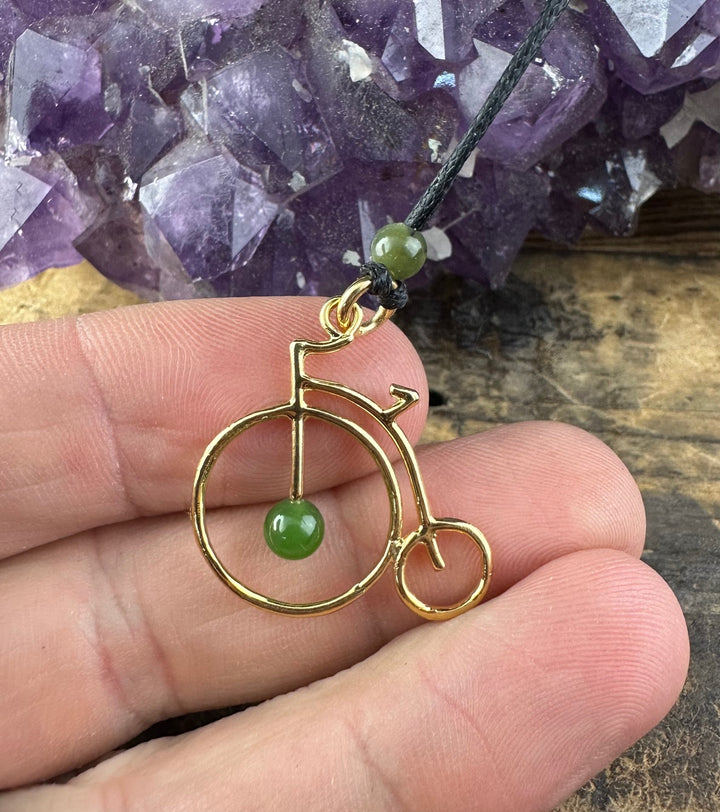 Handheld view of the gold penny farthing pendant with a green jade bead on a black cord near amethyst crystals.