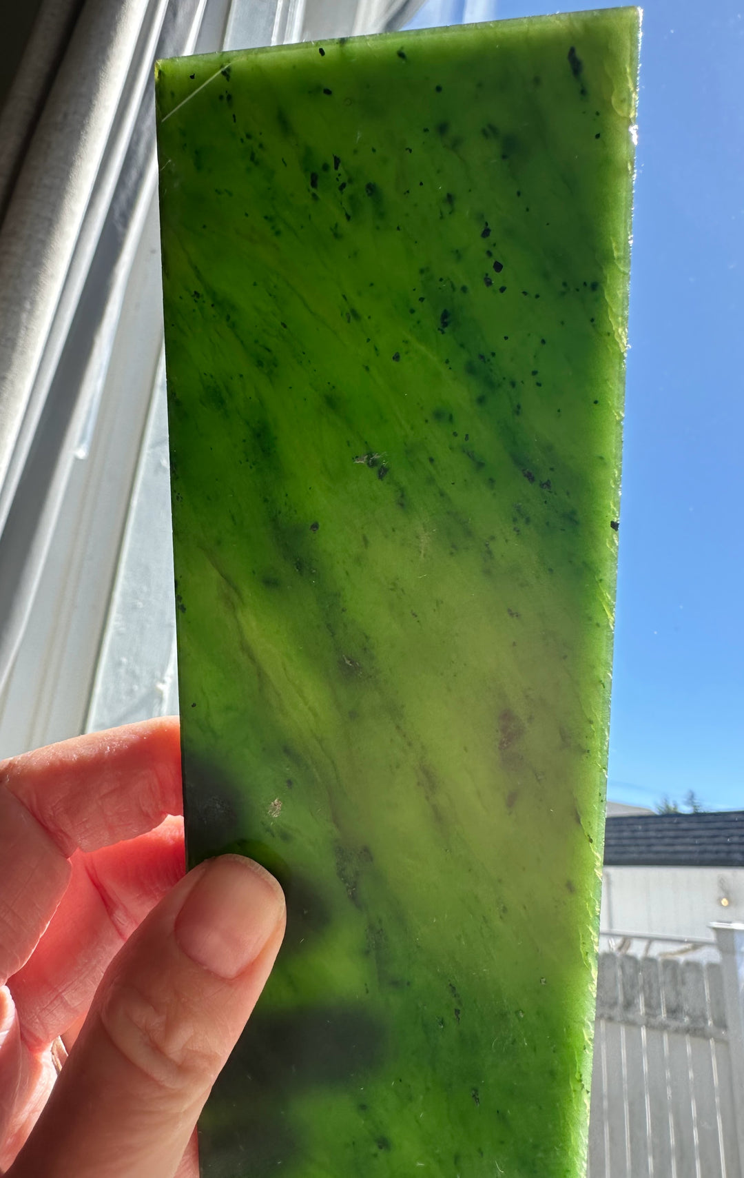 Bright green A Grade Canadian jade slab held upright by fingers against a window with dark speckles.