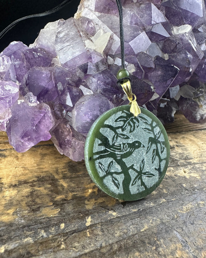 Round green jade pendant with a bird carving on a branch, suspended from a black cord beside purple amethyst crystals
