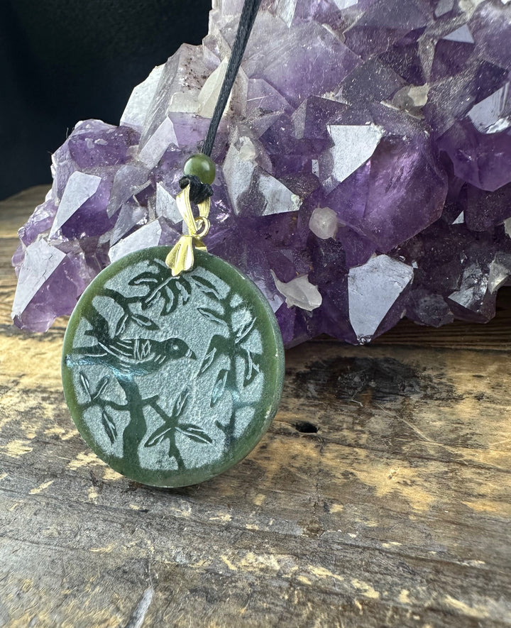 Green round jade pendant featuring a bird among branches resting on weathered wood with amethyst crystals in the background