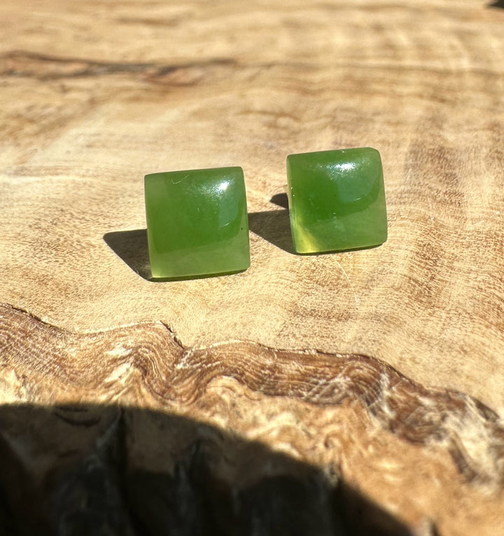 Two square jade earrings with bright green stones resting on a wooden surface