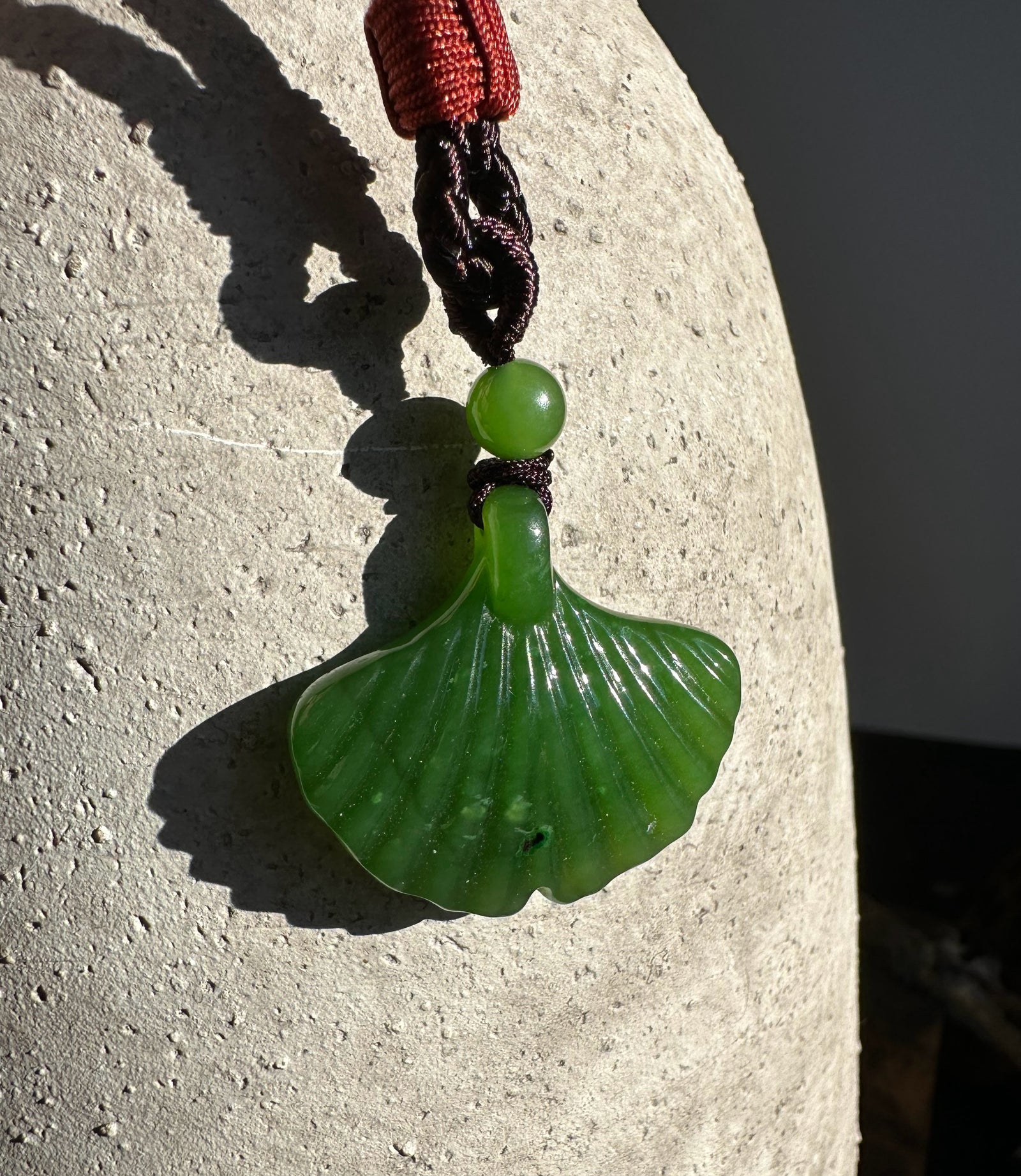 Green jade ginkgo leaf pendant 30mm on a braided cord against a textured stone background