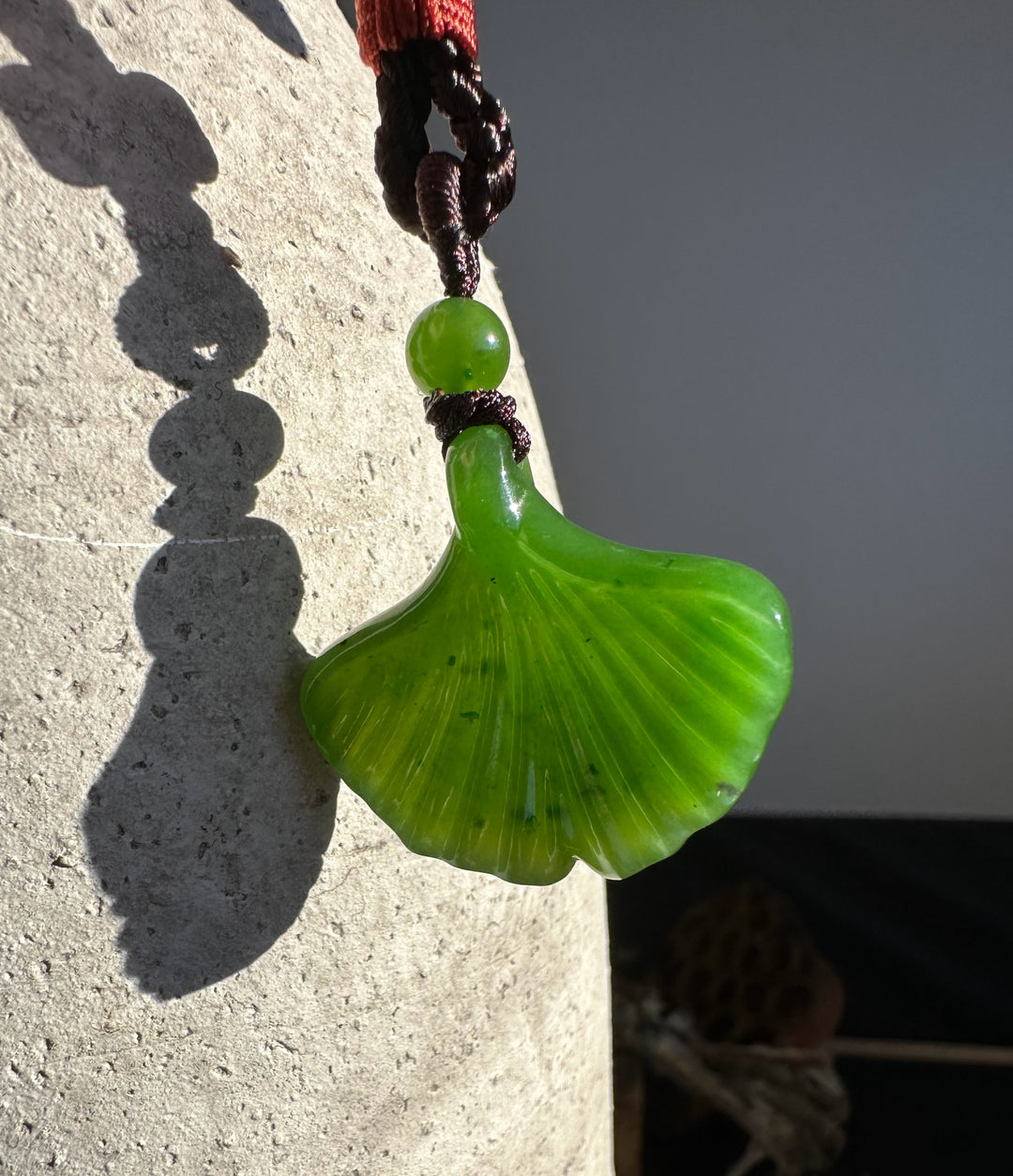 Canadian Jade Ginkgo Leaf Pendant 30mm with green bead on a dark braided cord hanging by a stone pillar