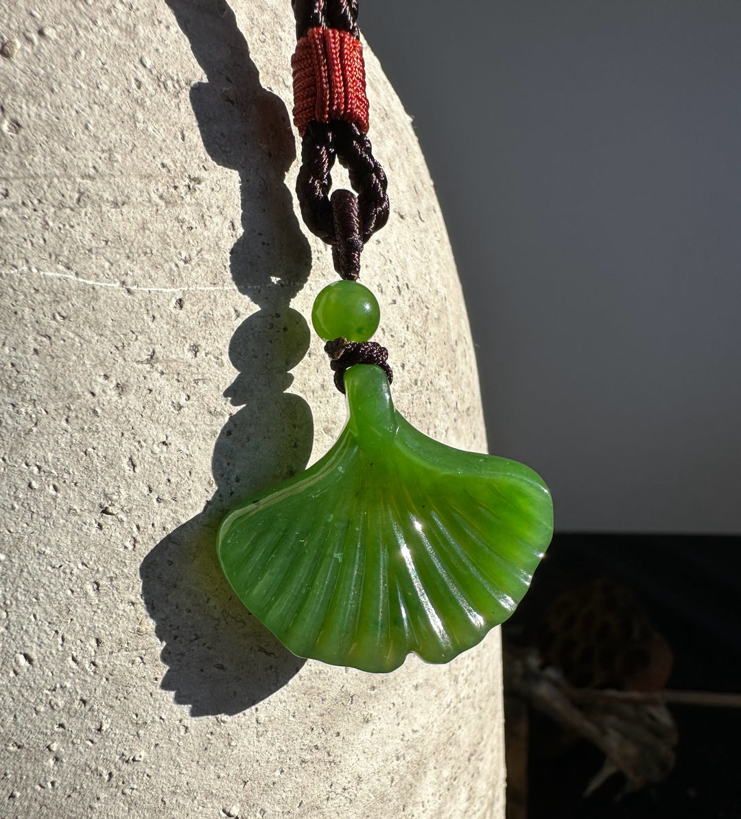 Close up of green jade ginkgo leaf pendant suspended from brown and red braided cord