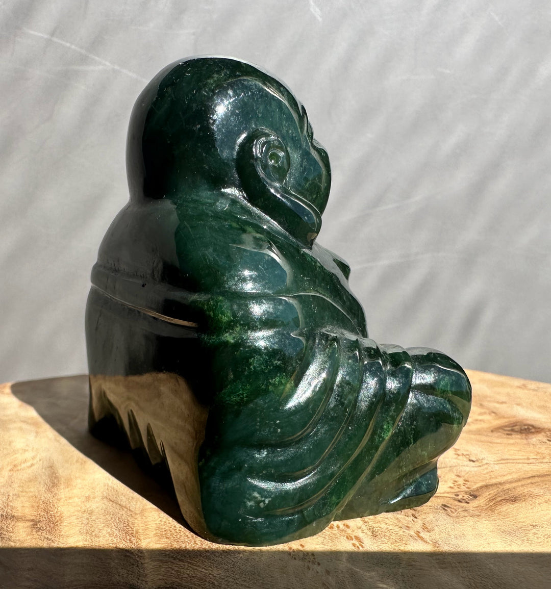 Side view of a 2.5 inch dark green Happy Jade Buddha seated with carved robes on a wooden surface