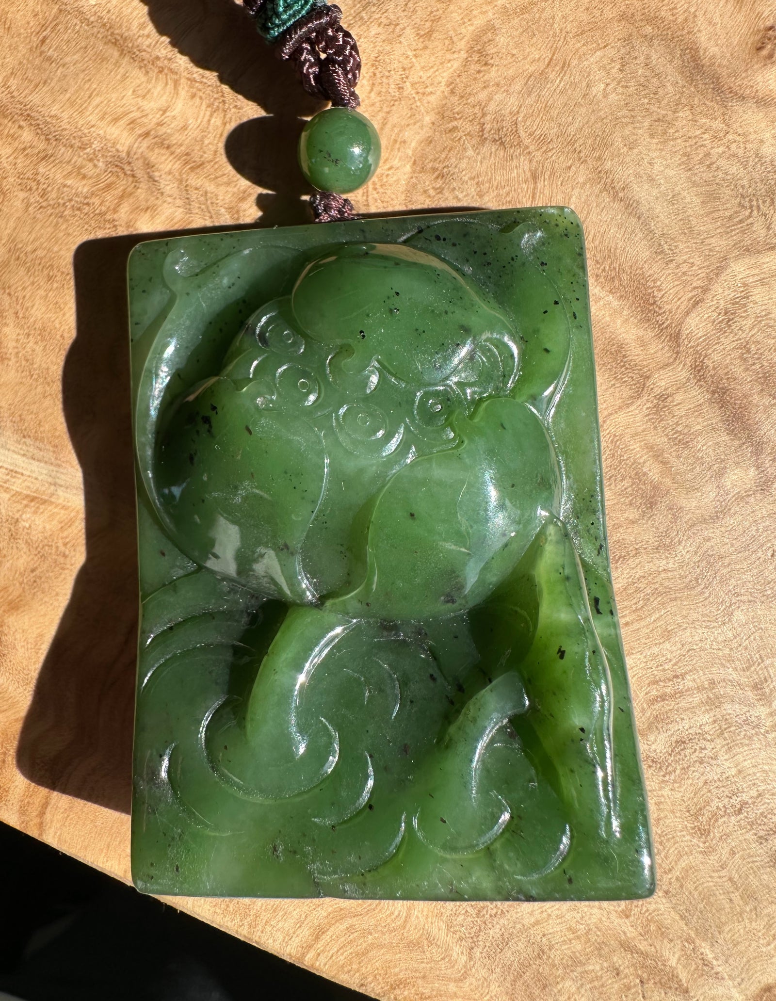 Green Canadian jade large lotus pendant carved with a swirling lotus on a rectangular slab, suspended by a braided cord.