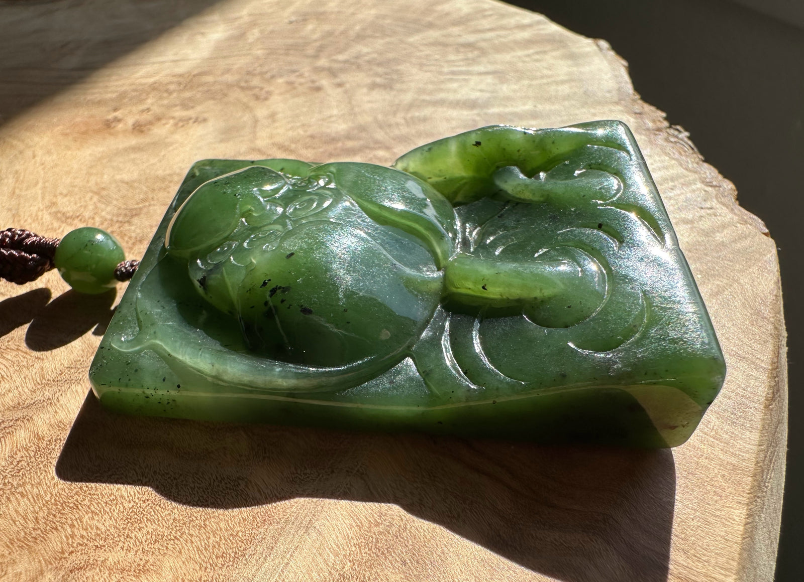 Green jade large lotus pendant resting on a wood surface, rectangular slab with etched petal lines and a braided cord.