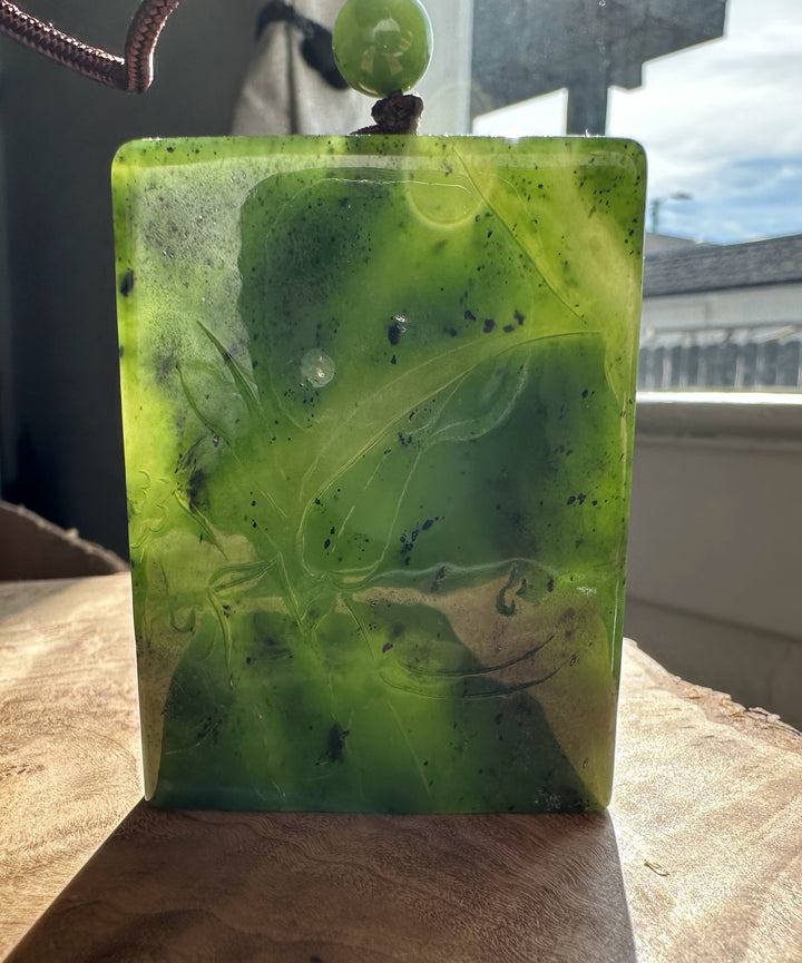 Rectangular green jade pendant featuring a lotus carving, suspended by a brown cord and green bead.