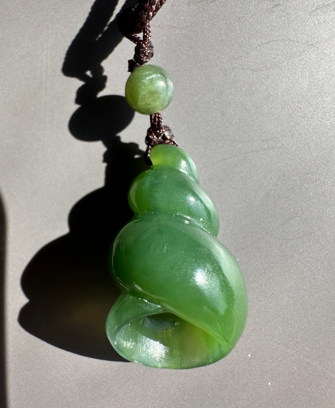 A green jade spiral pendant charm with a small round green bead and dark brown cord, photographed on a neutral gray background with a strong shadow.
