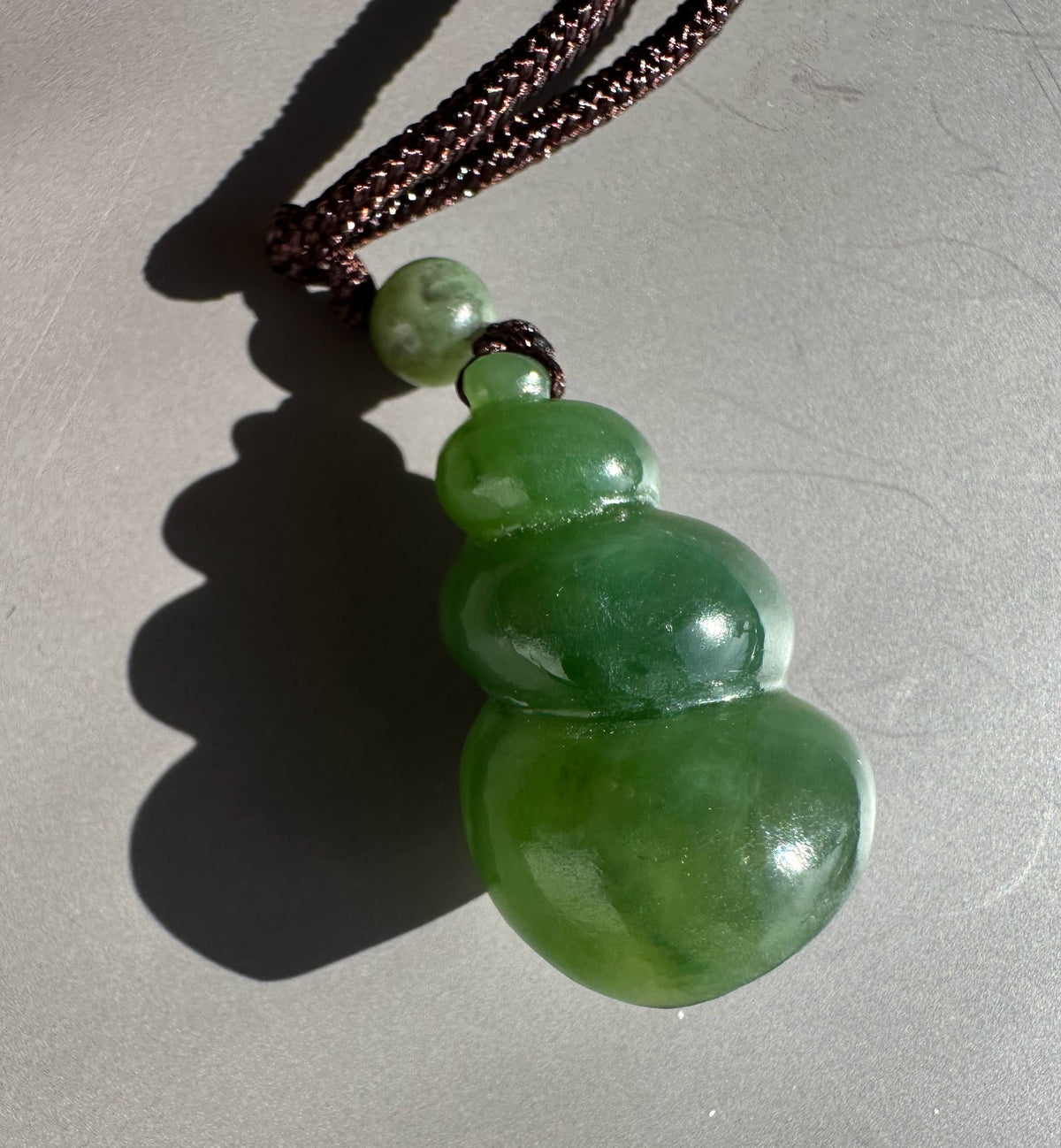 A green jade huggie pendant necklace on a dark brown braided cord, photographed on a neutral gray surface with a hard shadow.