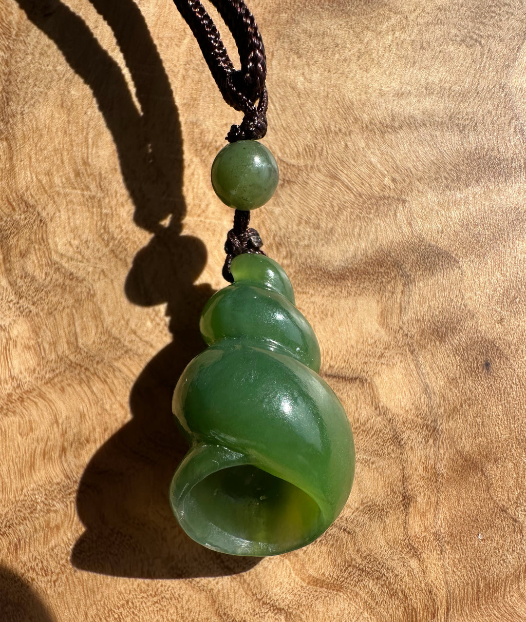 A green jade shell-shaped pendant necklace with a dark brown braided cord, displayed on a textured wooden surface.