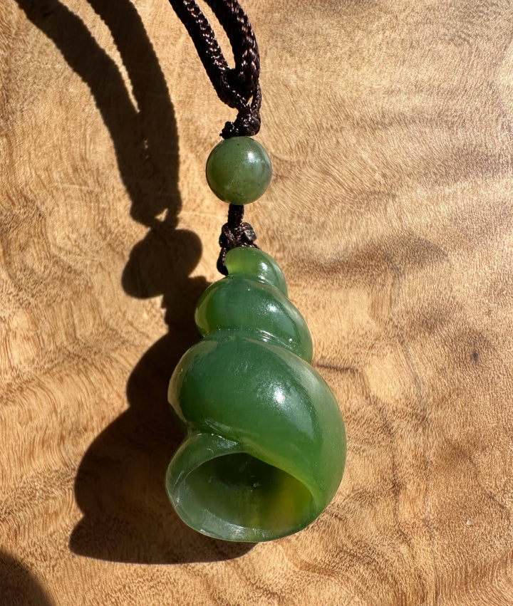 A green jade shell-shaped pendant necklace with a dark brown braided cord, displayed on a textured wooden surface.