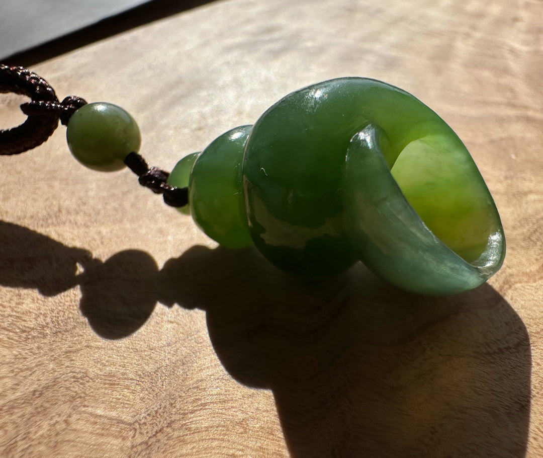 A polished green jade pendant with a carved spiral design, attached to a dark brown cord with a small green bead, resting on a wooden surface under direct sunlight.