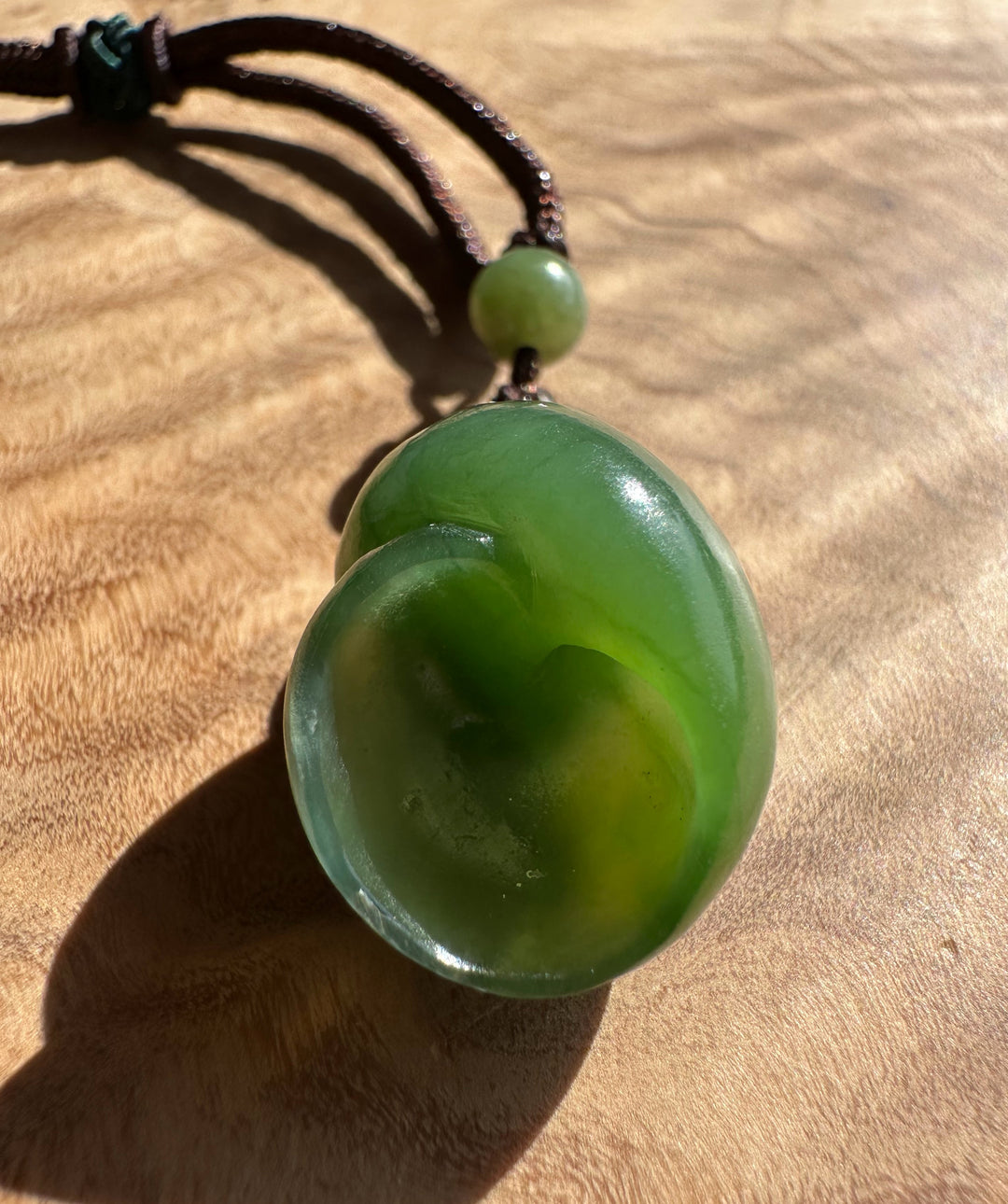 A carved green jade pendant on a dark brown cord, resting on a wooden surface under direct sunlight.
