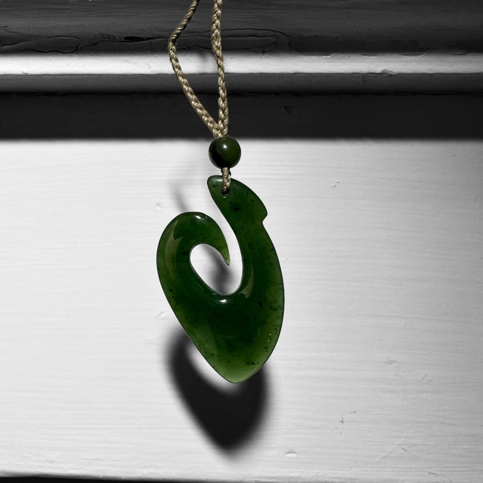 Green jade fish hook pendant on a tan braided cord with a small bead above