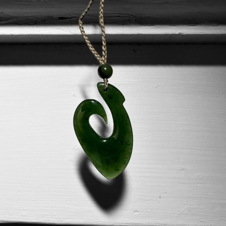 Green jade fish hook pendant on a tan braided cord with a small bead above