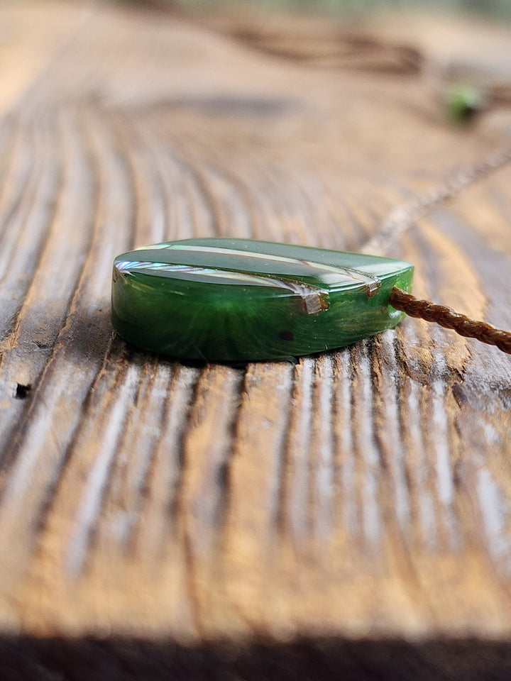 Side view of green jade teardrop pendant with abalone inlay on a braided cord resting on wood