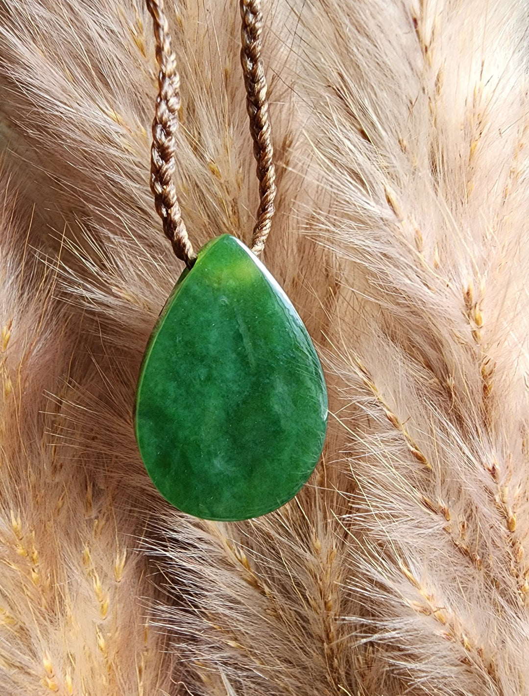 Teardrop nephrite jade pendant with abalone inlay hanging on a brown braided cord against dried grasses