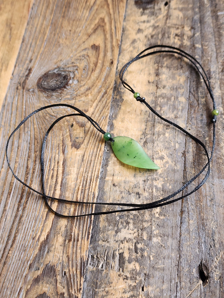 Jade leaf pendant on a black cord with a green bead resting on a rustic wooden surface
