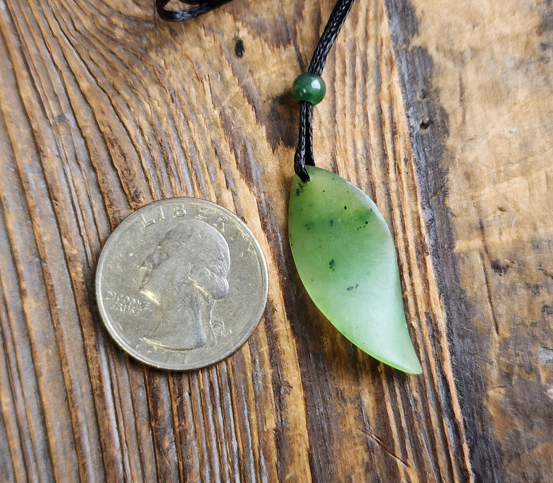 Green jade leaf pendant on black cord with small bead, placed next to a quarter for scale on a wood surface.