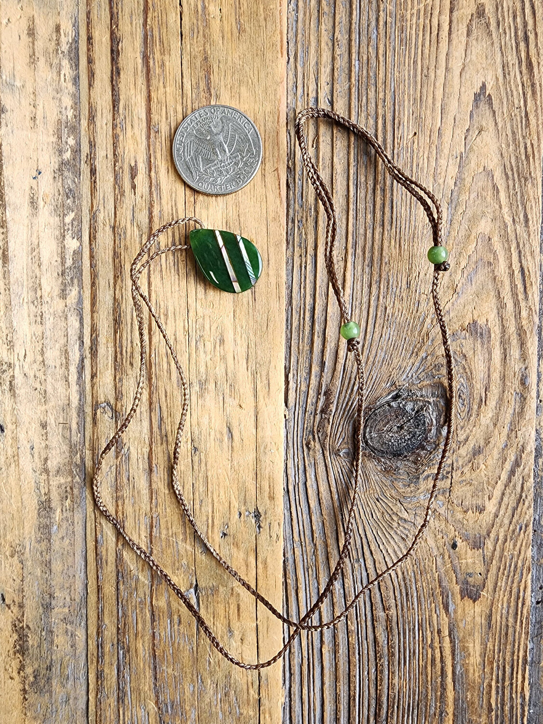 Teardrop nephrite jade pendant with abalone on a brown braided cord shown on a wooden surface with a quarter for scale