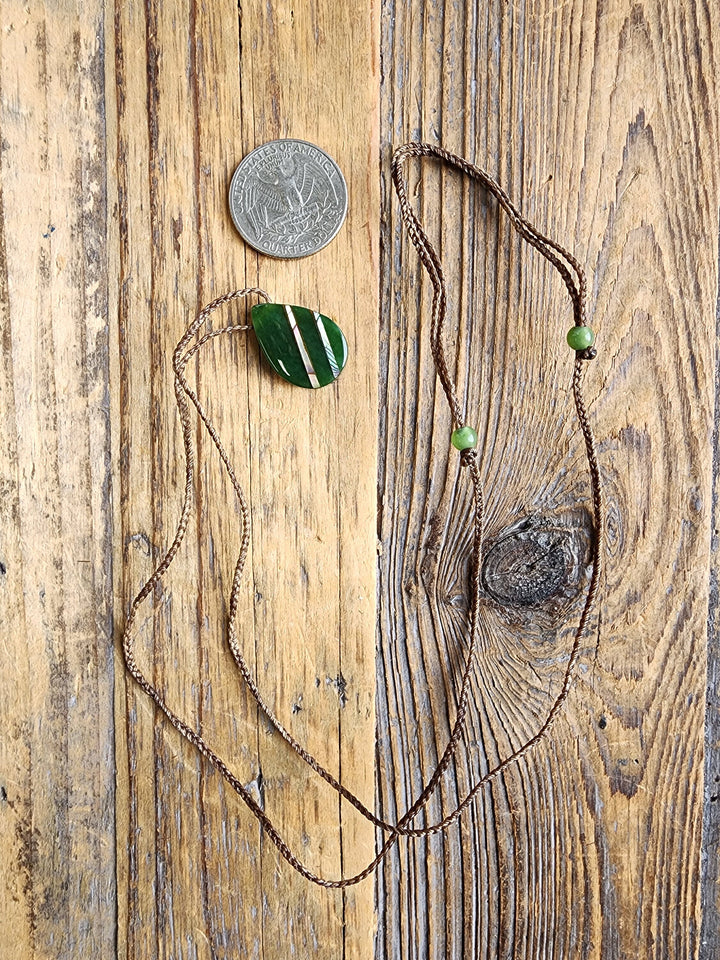 Teardrop nephrite jade pendant with abalone on a brown braided cord shown on a wooden surface with a quarter for scale