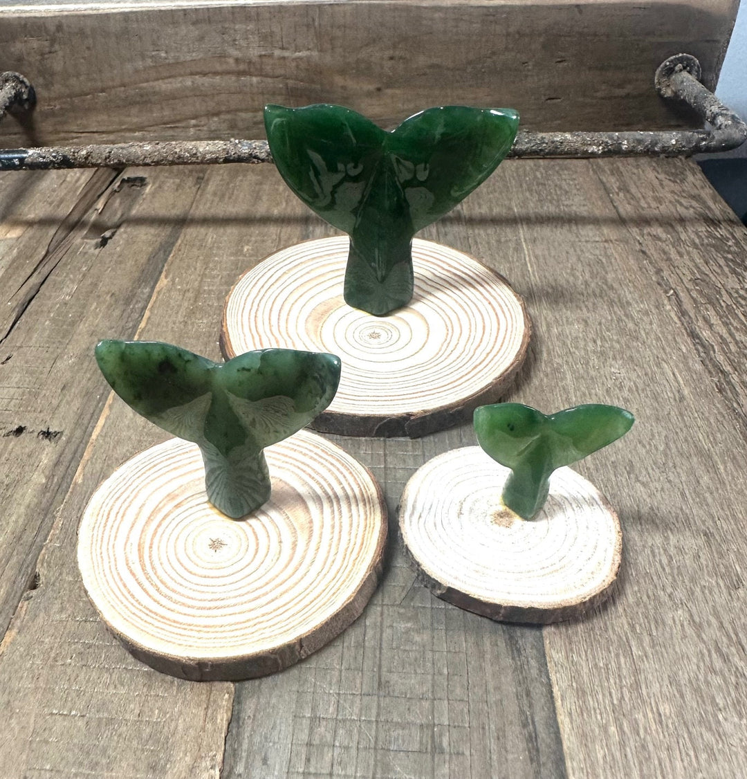 Three green jade whale tail sculptures on round wood bases of varying sizes arranged on a rustic wooden surface.