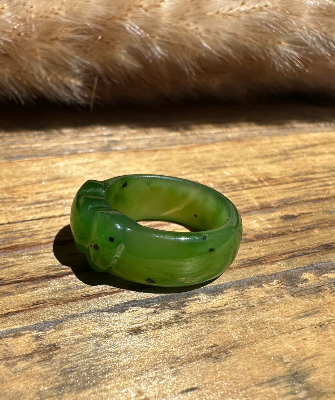 A carved green stone ring with a small carved figure on the band, resting on a rustic wooden surface with a soft beige fur texture in the background.