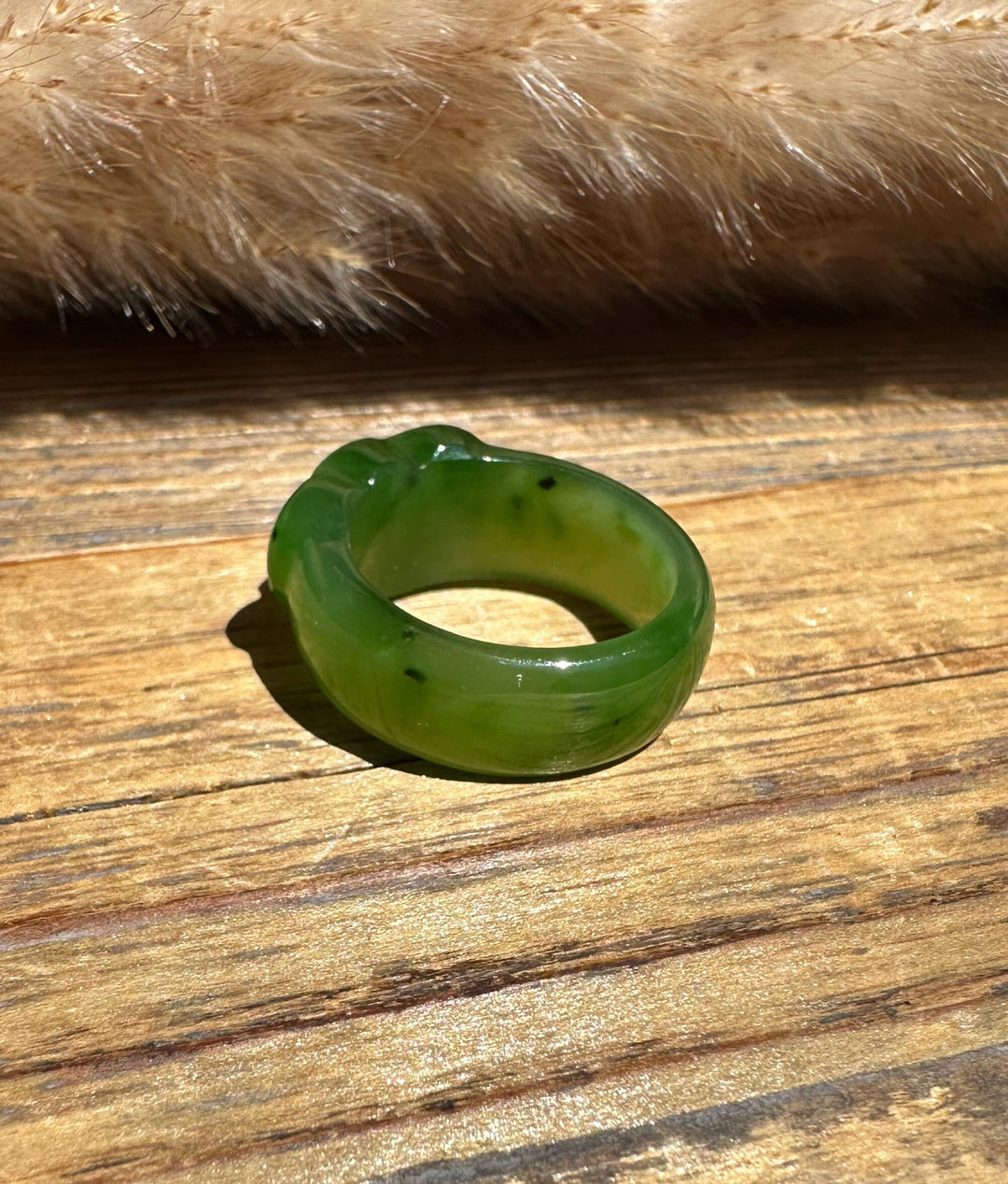 A carved green jade ring, resting on a rustic wooden surface with a soft beige faux fur prop in the background.