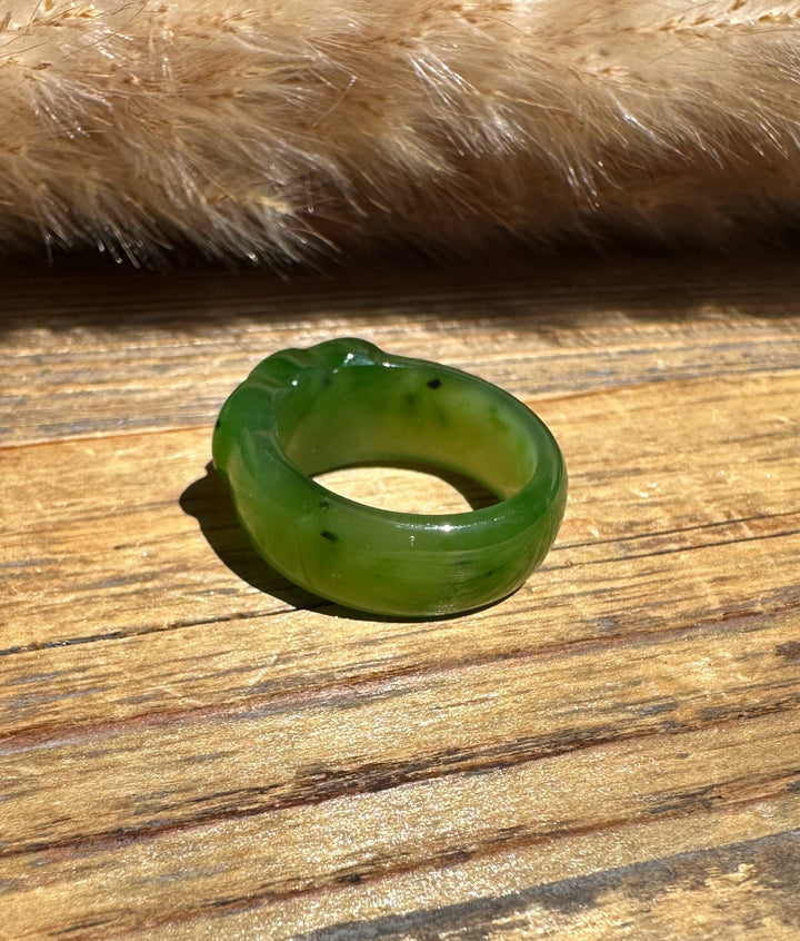 A carved green jade ring, resting on a rustic wooden surface with a soft beige faux fur prop in the background.