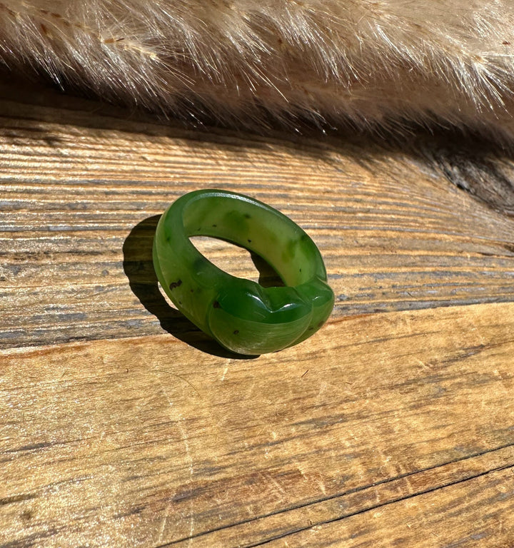 A carved green jade ring with a textured, organic shape, resting on a rustic wooden surface with a fur pelt in the background.