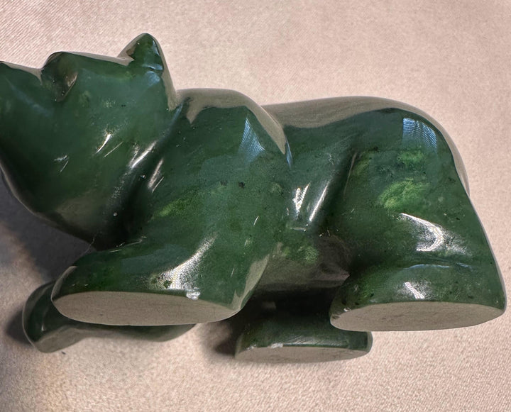 A polished dark green jade figurine of a resting animal, likely a bear or dog, shown on a neutral beige fabric background.