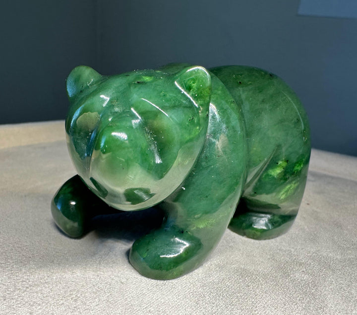 A polished green jade bear figurine, carved with a glossy finish, resting on a light beige fabric surface against a dark gray background.