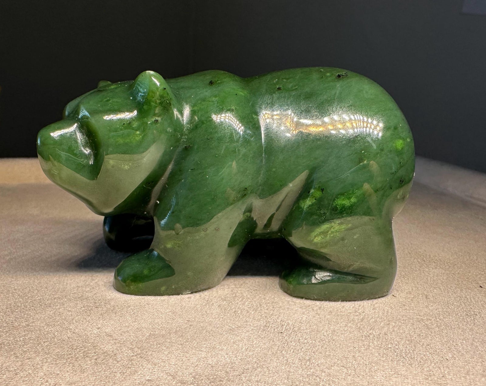 A polished green jade  bear figurine carved with natural stone inclusions, standing on a beige fabric surface against a dark gray background.