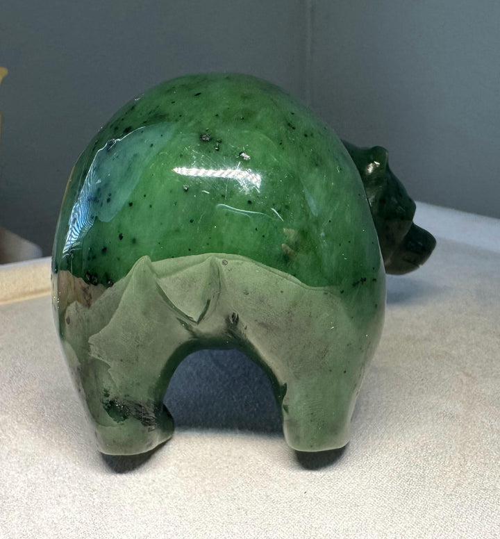 A green jade stylized animal with a rounded back, short legs, and a small head on the right, photographed on a light fabric surface against a gray background.