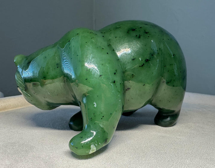 A glossy green jade bear figurine with dark inclusions, standing on a light beige surface against a gray background.