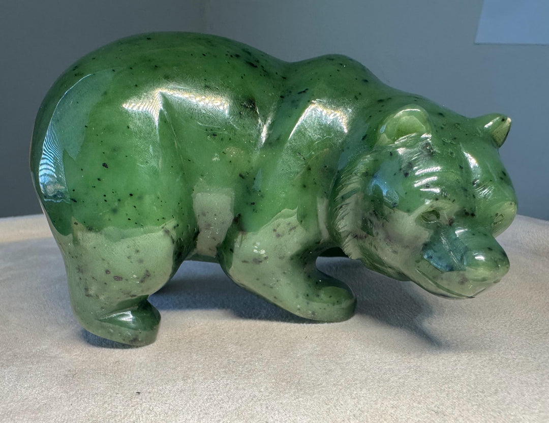 A polished green jade bear figurine with natural speckles, standing on a light beige fabric surface against a neutral gray background.