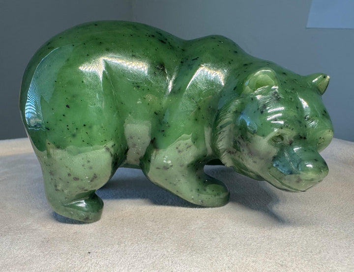 A polished green jade bear figurine with natural speckles, standing on a light beige fabric surface against a neutral gray background.