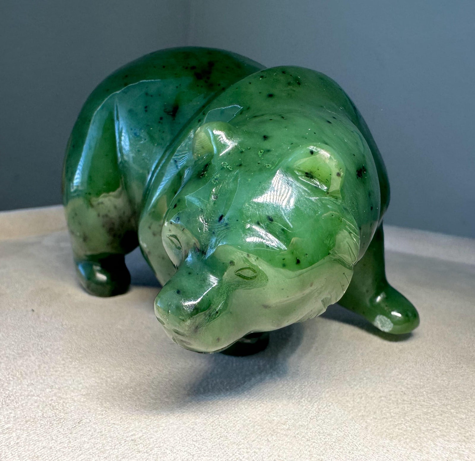 A polished green jade polar bear figurine with natural dark inclusions, displayed on a light beige surface against a gray background.