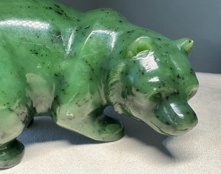 A polished green jade bear figurine with natural dark inclusions, shown in a close-up studio shot against a neutral gray background.