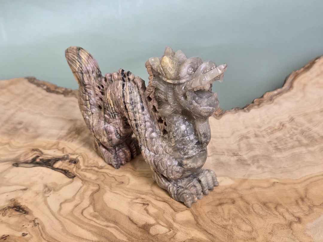 A small carved dragon figurine made from textured gray stone or resin, displayed on a natural wood slab with a pale blue-green background.