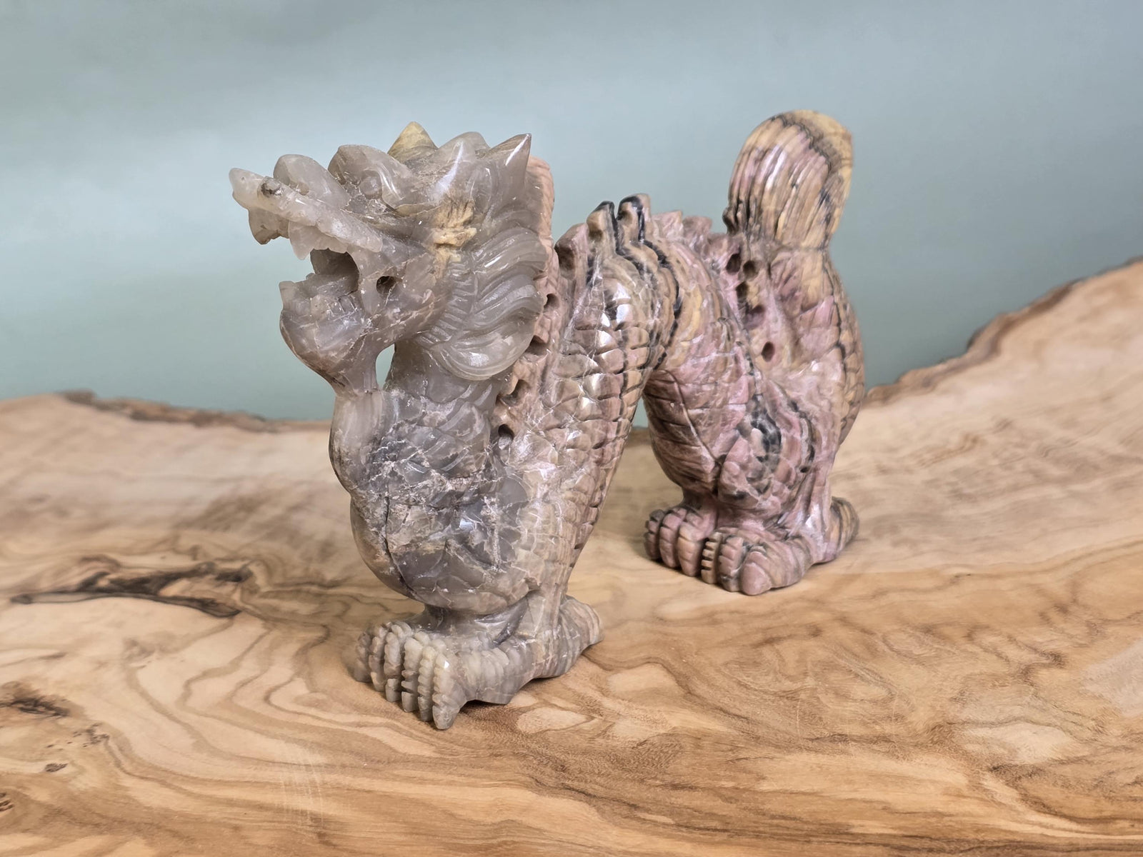 A small carved stone dragon figurine with a gray and pinkish stone texture, displayed on a natural wood slice against a soft blue-gray background.