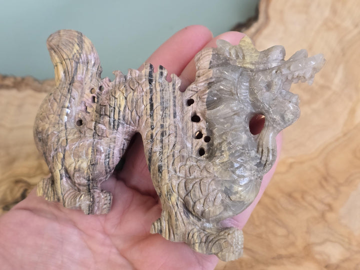 A hand holding a carved natural wood object with a dragon-like figure, featuring a small round reddish-brown bead or stone set into its side, against a blurred background.