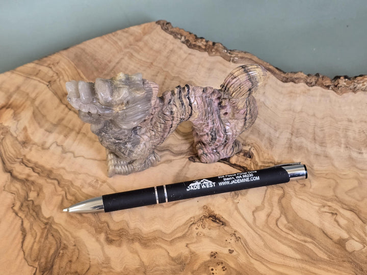 A black Jades West ballpoint pen with silver accents lies on a polished wood slab next to a carved stone figurine, with a muted green wall in the background.